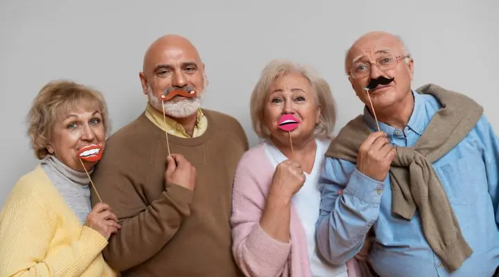 Cuatro miembros de la generación del baby boom están juntos, sonriendo y sosteniendo divertidos adornos de papel con forma de labios y bigotes frente a sus caras sobre un fondo liso Early Boomers.
