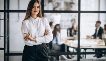 Una mujer de negocios que utiliza las relaciones públicas como herramienta para mejorar las ventas, parada con confianza frente a una pared de vidrio en una oficina.