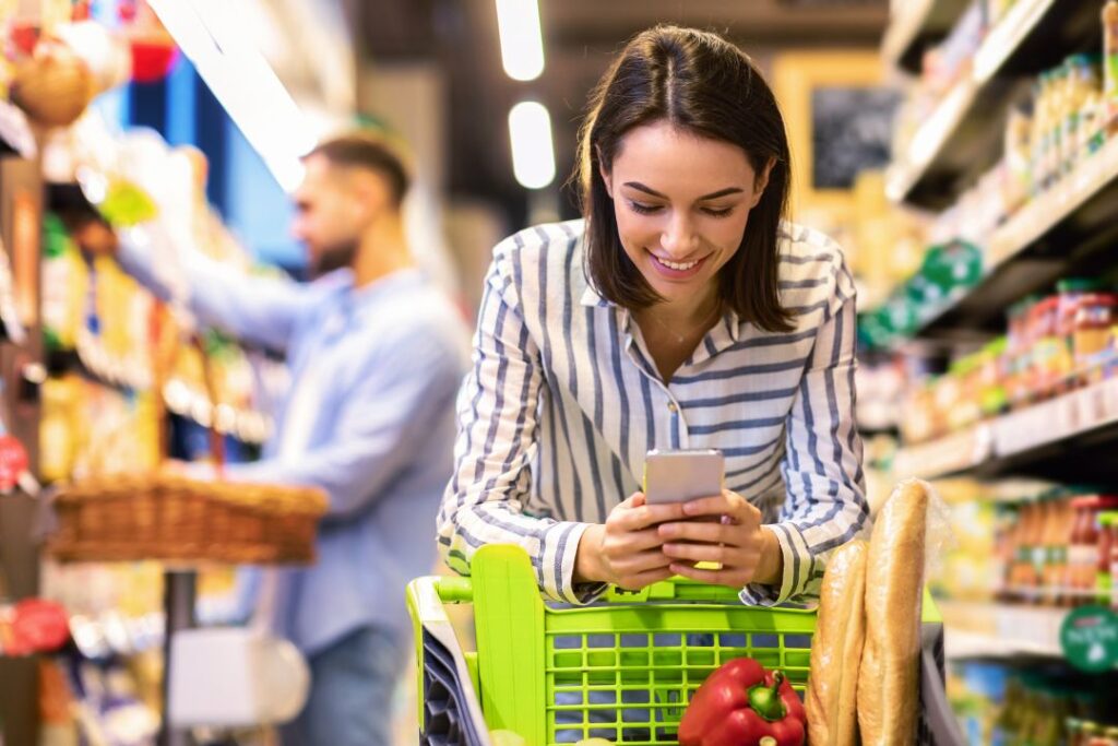 Una mujer navega con su teléfono mientras compra en un supermercado, potencialmente interactuando con marketing de aplicaciones..