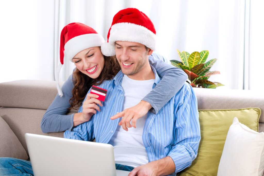 Una pareja con gorros de Papá Noel celebrando la temporada navideña, sentada en un sofá y usando una computadora portátil.