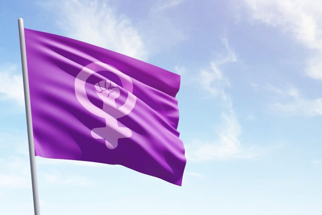 Una bandera morada que presenta una combinación del símbolo del género femenino y un puño en alto ondeando contra un cielo azul parcialmente nublado, conmemorando el  8M Día de la Mujer.