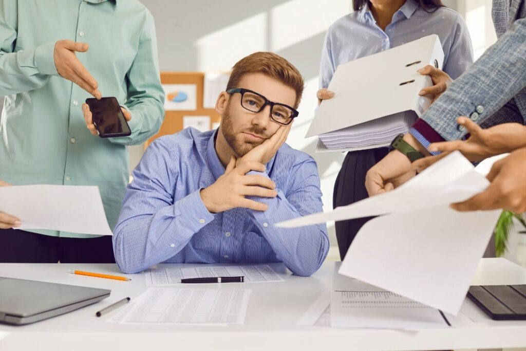 Un hombre con gafas reflexionando en un escritorio rodeado de varias personas entregándole papeles y un teléfono inteligente durante sus descansos laborales.