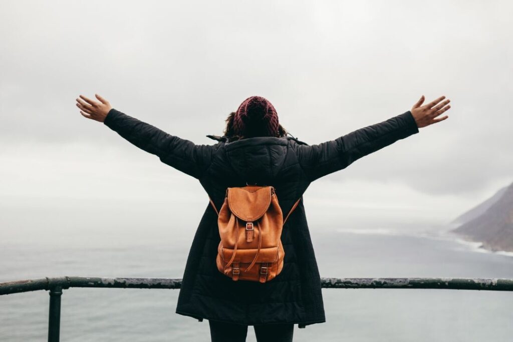 Una mujer con una mochila parada sobre un acantilado con vista al océano con los brazos extendidos, disfrutando de experiencias personalizadas.