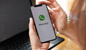 Una persona sosteniendo un teléfono inteligente con el logo de WhatsApp Empresarial en la pantalla, sentada en un escritorio con una computadora portátil.