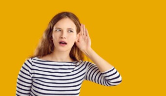 Mujer joven con una camisa a rayas, llevándose la mano a la oreja, escuchando historias atentamente, sobre un fondo amarillo.