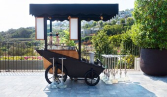 Un carrito de bebidas móvil con un dosel negro está instalado al aire libre cerca de una barandilla. El carrito está equipado con dispensadores y rodeado de vegetación y edificios en la ladera al fondo, creando uno de esos espacios perfectos instagrameables.