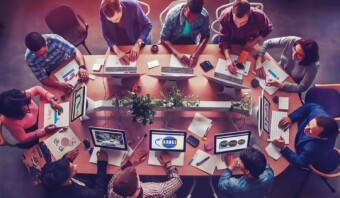 Un grupo de personas sentadas alrededor de una mesa en una oficina, colaborando y trabajando en computadoras portátiles. Entre los papeles y las plantas esparcidas sobre la mesa, las discusiones sobre la integración de la IA en su proyecto bullen de entusiasmo.