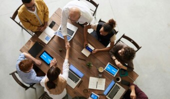 Un grupo de personas sentadas alrededor de una mesa de madera con ordenadores portátiles y portátiles, encarnando una empresa inclusiva. Dos personas en el centro se dan la mano.