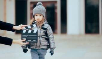 Un niño pequeño, vestido con un abrigo de invierno, gorro y guantes, se encuentra al aire libre con una claqueta preparada para el rodaje de una película. La escena captura la presencia encantadora que se ve a menudo en niños en publicidad.