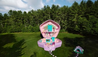 Una casa de muñecas grande y abierta con varias habitaciones y una decoración de temática rosa, que recuerda a un juego de Polly Pocket de tamaño real, está situada sobre césped verde rodeada de árboles densos. Una pequeña estructura similar a una tienda de campaña está ubicada a la derecha de la casa de muñecas.