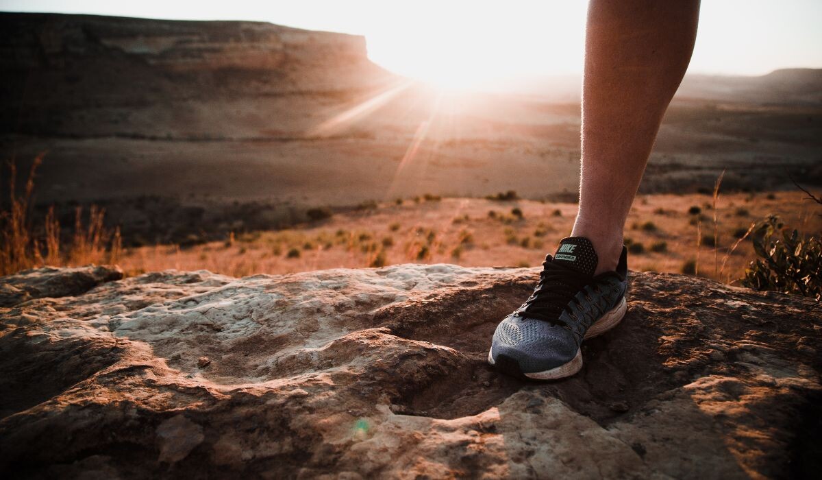Una persona que lleva zapatillas para correr se encuentra en un terreno rocoso con un paisaje desértico y una puesta de sol de fondo.