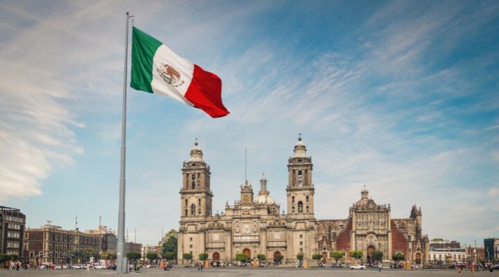 Una gran bandera mexicana, uno de los símbolos patrios de la nación, ondea frente a la histórica Catedral Metropolitana en la plaza principal de la Ciudad de México en un día claro.