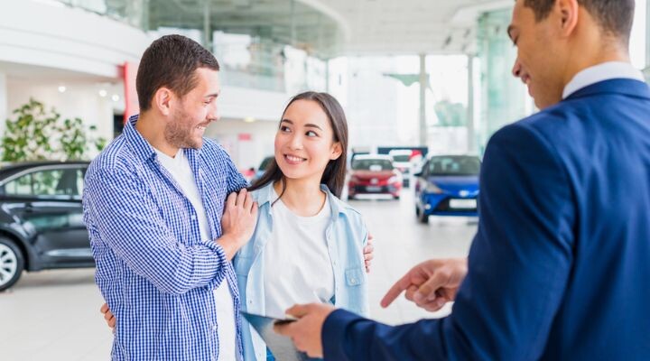 Una pareja sonriente conversa con un vendedor en una sala de exposición de automóviles sobre las comisiones de venta. El hombre rodea a la mujer con el brazo mientras se encuentran cerca de varios automóviles estacionados.