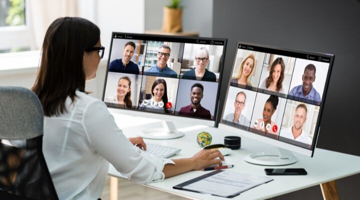 Una persona con camisa blanca participa en una videoconferencia en dos monitores de computadora, cada uno de los cuales muestra a varios participantes en ventanas separadas de comunidades online.