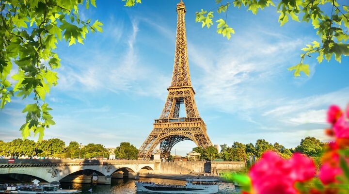 La Torre Eiffel de París, vista desde la orilla de un río cercano, con hojas verdes y flores rosas en primer plano en un día soleado, evoca escenas sacadas directamente de "Emily in París".