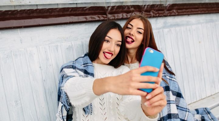 Dos mujeres se toman una selfie mientras sacan la lengua, ambas vistiendo suéteres blancos y envueltas en una manta a cuadros azules y blancos, listas para compartir su momento divertido en las redes sociales.