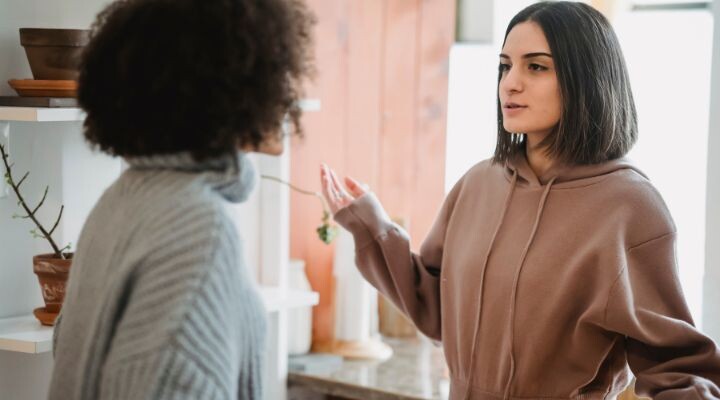 Dos mujeres mantienen una conversación seria en una habitación con estanterías y macetas que recuerdan escenas de reality shows. Una lleva una sudadera marrón y la otra un jersey gris.