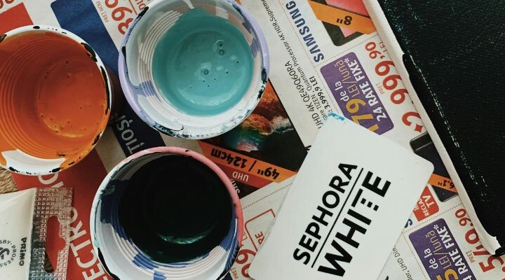 Vasos de plástico con pintura naranja, azul y negra se encuentran junto a una tarjeta Sephora White sobre una superficie cubierta con papeles publicitarios; cada pieza contribuye a una escena narrativa colorida del storytelling.