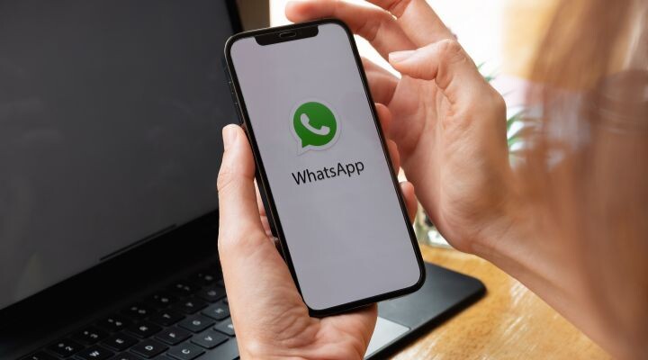 Una persona sostiene un teléfono inteligente que muestra el logotipo de WhatsApp Business en la pantalla, con una computadora portátil abierta en el fondo.