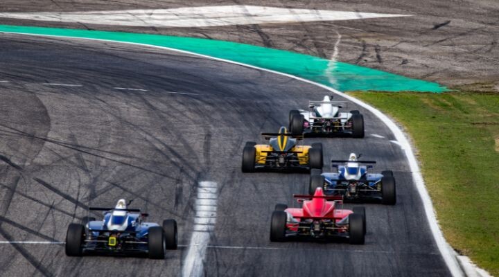 Cinco coches de carreras de Fórmula 1, cada uno de un color diferente, corren a toda velocidad por una pista con curvas, dejando marcas de neumáticos en el asfalto.