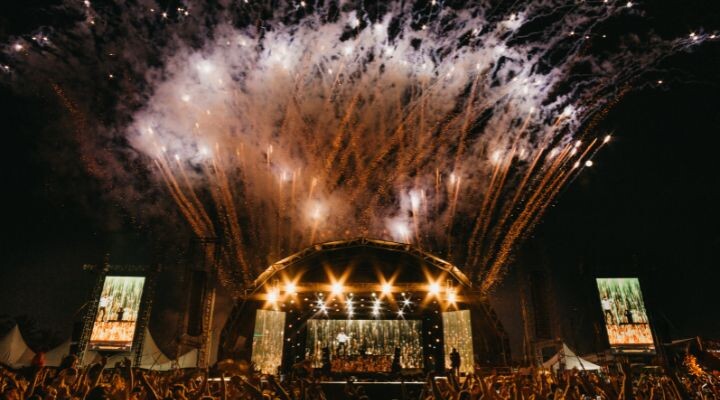 Un vibrante concierto al aire libre que reúne a una gran multitud frente a un escenario iluminado donde Luis Miguel deslumbra bajo el cielo nocturno. Los fuegos artificiales explotan en el cielo, mientras dos grandes pantallas flanquean el escenario, amplificando la atmósfera electrizante.