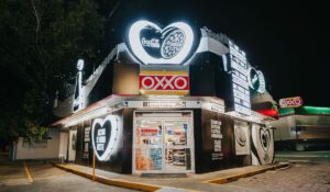 Vista nocturna de una tienda de conveniencia OXXO iluminada con señalización y anuncios de marca, incluido un gran logotipo de Coca-Cola y decoraciones de corazones.