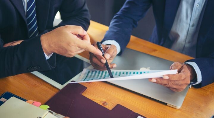 Dos personas con traje están sentadas en una mesa de madera y discuten sobre un documento con un gráfico. Uno señala mientras el otro sostiene el documento sobre blindaje corporativo. También hay una computadora portátil y carpetas sobre la mesa, lo que enfatiza su enfoque en el blindaje corporativo.