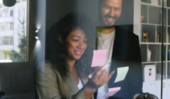 Dos personas, sonrientes mientras participan en una efectiva lluvia de ideas, anotan ideas en notas adhesivas fijadas a una pared de vidrio en un entorno de oficina.