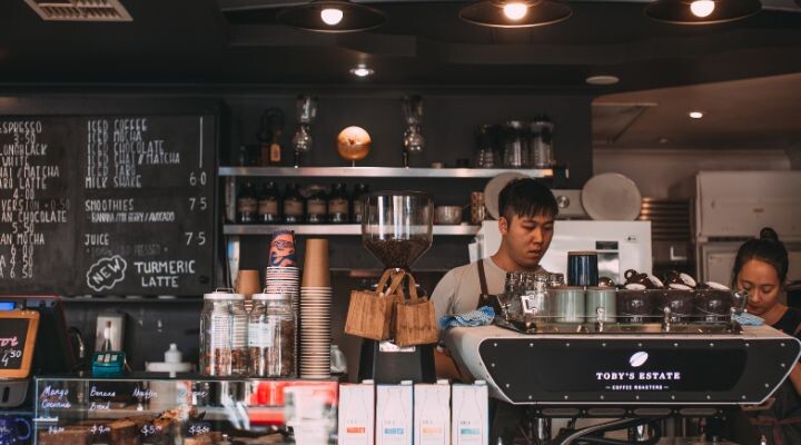 En una cafeterías bulliciosa, un barista interactúa con un cliente en medio de una variedad de elementos, como el tablero del menú, las tazas de café y la siempre activa máquina de café.