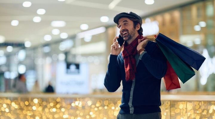 Un hombre se encuentra en un centro comercial, sonriendo y hablando por teléfono, discutiendo su estrategia de medio ambiente para fin de año, mientras sostiene coloridas bolsas de compras sobre su hombro.