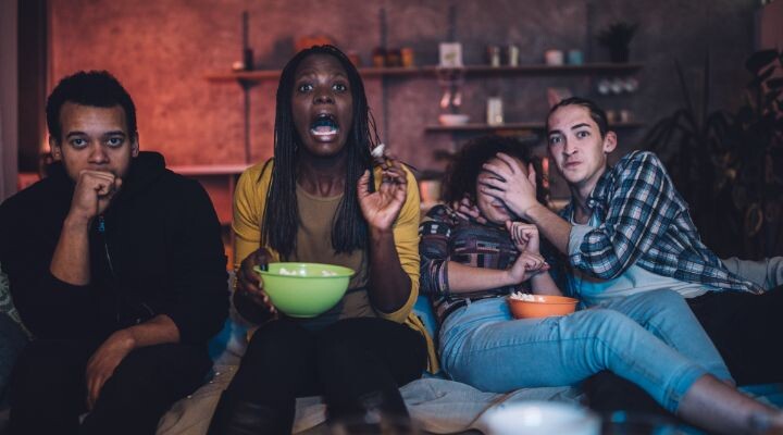 Cuatro personas se sientan en un sofá, viendo una película de Halloween con expresiones de sorpresa y anticipación, mientras sostienen cuencos de bocadillos.