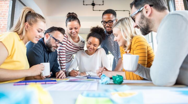 Un grupo diverso de personas se sienta y se para alrededor de una mesa, colaborando en la idea y sonriendo, con papeles y bebidas frente a ellos.