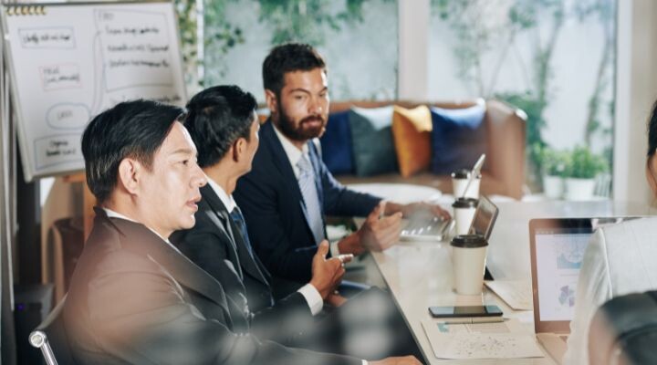 Cuatro personas vestidas de negocios están en una reunión, discutiendo estrategias de relaciones públicas alrededor de una mesa llena de computadoras portátiles y papeles. Una pizarra con notas es visible en el fondo, lo que refleja sus esfuerzos de colaboración.