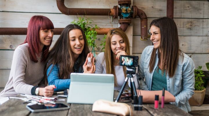 Cuatro mujeres se sientan en una mesa con productos de maquillaje, una computadora portátil y una cámara, aparentemente comprometidas y sonrientes. Parecen estar grabando un video o realizando una transmisión en vivo, encarnando la esencia de los influencers.