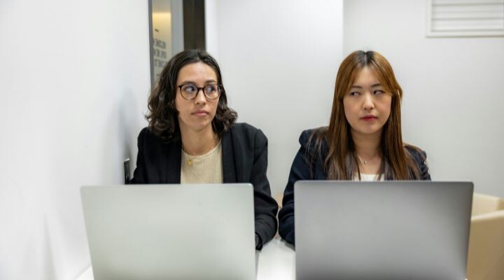 Dos personas están sentadas a una mesa, cada una trabajando con un ordenador portátil, con la atención centrada en ellas. Vestidas con ropa de trabajo, parecen estar debatiendo políticas de trabajo destinadas a mejorar la productividad y la colaboración. politicas en el trabajo,