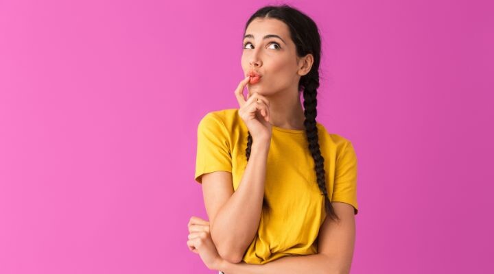 Una persona con cabello trenzado y una camisa amarilla se encuentra sobre un fondo rosa, contemplando su marca personal con un dedo en los labios.