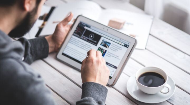 Una persona utiliza una tableta con un lápiz óptico en una mesa de madera blanca, tal vez navegando en redes sociales. Cerca hay una taza de café y un libro abierto.