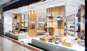 Exhibición en escaparate con diversos bolsos y accesorios en estantes y plataformas detrás de una gran ventana de vidrio en un espacio comercial bien iluminado.