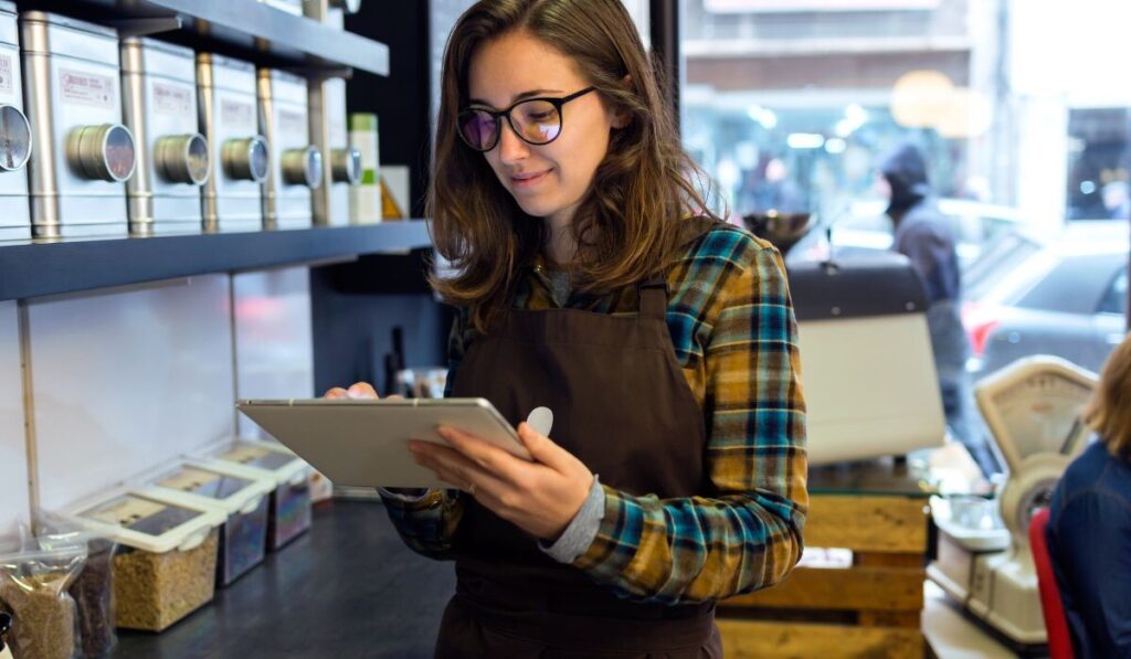 Una mujer con gafas y delantal usa una tableta en una cafetería, rodeada de recipientes y equipos de café. retail media 