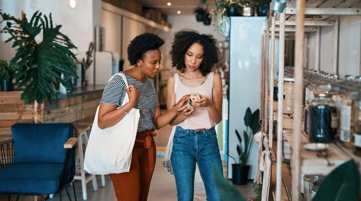 Dos mujeres examinan juntas un frasco en una tienda, atraídas por su contenido. Una lleva una bolsa de mano mientras participan en una segmentación emocional, en la que hablan de las emociones asociadas a cada producto. La tienda cuenta con plantas exuberantes y estanterías repletas de artículos diversos que captan su interés.