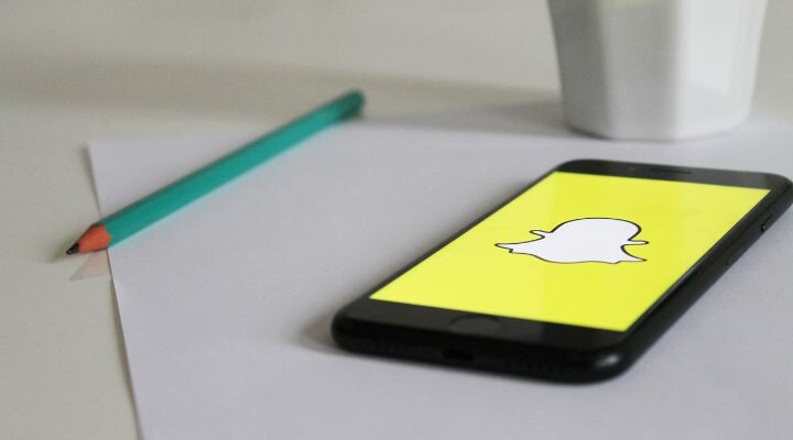 Un teléfono inteligente con el logotipo de Snapchat reposa sobre un papel blanco, acompañado por un lápiz verde azulado y una taza blanca impecable.