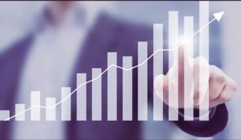 Una persona con traje señala un gráfico de barras ascendente con una superposición de un gráfico de líneas, lo que indica crecimiento o una tendencia positiva.