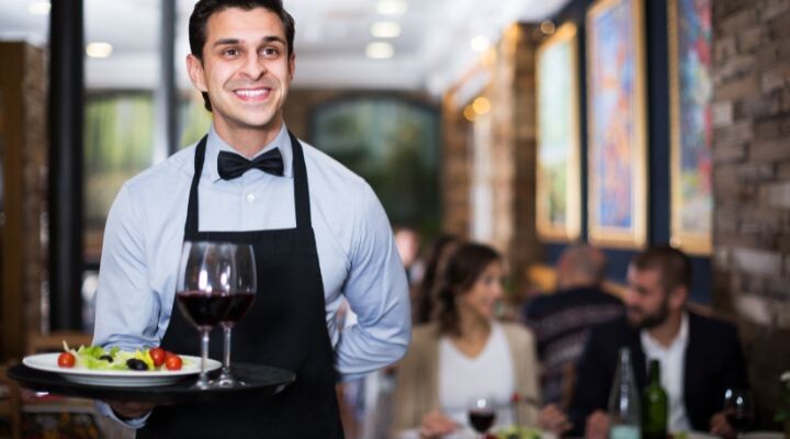 Un camarero con delantal negro y pajarita sirve una bandeja con vino y ensalada en un restaurante. Al fondo hay comensales sentados en mesas. storytelling 