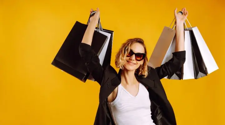 Una persona con gafas de sol y una chaqueta negra sostiene triunfante varias bolsas de compras en el aire sobre un fondo amarillo brillante, encarnando el espíritu del Buen Fin.