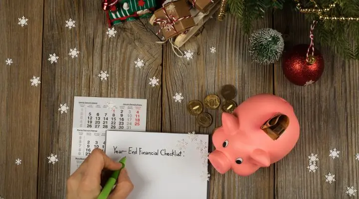 Una persona escribe una "Lista de control financiero de fin de año" en un bloc de notas. Cerca de allí, entre las decoraciones navideñas y los regalos envueltos, hay una alcancía, monedas y un calendario: un recordatorio de las compras de fin de año sobre la superficie de madera.