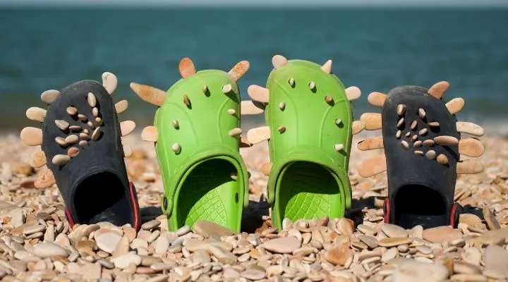 Dos pares de sandalias Crocs adornadas con conchas marinas se encuentran en posición vertical sobre los guijarros de una playa, con el mar extendiéndose pacíficamente en el fondo.