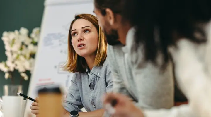 Una mujer con una camisa gris habla durante una reunión, sosteniendo un bolígrafo con un aire de autoridad común entre las emprendedoras mexicanas. Un hombre escucha atentamente y, al fondo, se ve un rotafolio lleno de ideas.