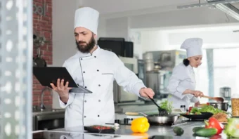 En una cocina llena de actividad, dos chefs elaboran delicias culinarias. Uno equilibra hábilmente las estrategias de marketing gastronómico en una computadora portátil mientras atiende una sartén chisporroteante, mientras el otro prepara con pasión los ingredientes frescos en la mesada, asegurándose de que cada plato cuente su propia historia deliciosa.