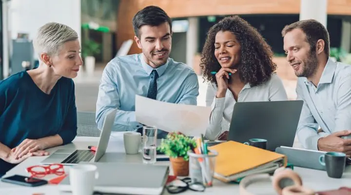 Un grupo de cuatro personas se sienta a una mesa y analiza una hoja de papel de la improvisación. Hay computadoras portátiles, cuadernos y tazas de café esparcidas por todas partes, creando una atmósfera alimentada por la creatividad espontánea y la colaboración.