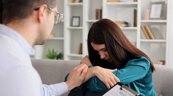 Una persona con anteojos sostiene suavemente la mano de una mujer sentada en un sofá, con la cabeza apoyada sobre los brazos cruzados. Estantes con libros y adornos enmarcan la escena, mientras navegan juntas por los desafíos de la salud mental en la búsqueda de empleo.